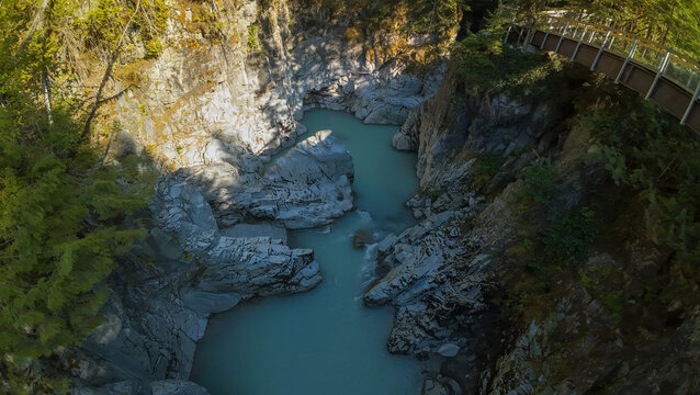 A high angle wide shot looks down a steep rock cliff canyon where the milky turquoise Mamquam River flows beneath a wooden boardwalk pedestrian path surrounded by dense forest trees.