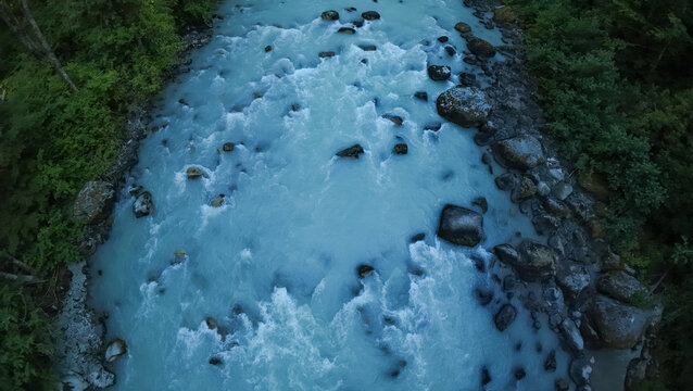 A top down aerial wide shot looks straight down at churning white water rapids and rocky boulders inside a fast flowing milky turquoise river flanked by dense woods.