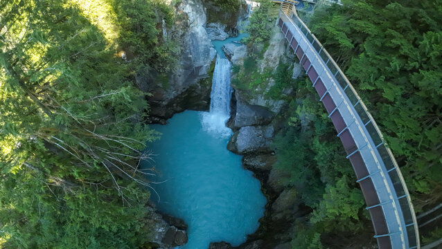 A high angle wide shot captures a curved wooden pedestrian boardwalk bridge hanging over a deep rocky canyon, revealing Mamquam Falls plunging into a turquoise river pool below.