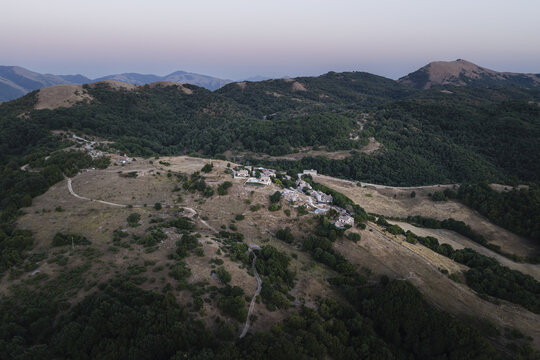 Aerial view of a small historic hamlet nestled on a hilltop surrounded by rolling green mountains and dense forests in the rural landscape of Spoleto, Umbria, Italy.