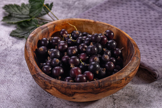 Black currants in rustic wooden bowl with green leaf on textured gray surface