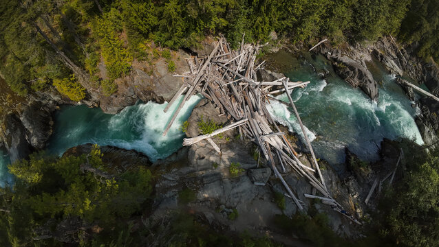 A top down close up aerial shot captures an extensive log jam of weathered driftwood logs blocked between large canyon boulders across the rushing turquoise Cheakamus River.