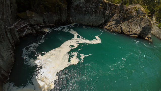 A high angle wide shot looks straight down at a patch of thick white water foam swirling on a calm deep turquoise river pool enclosed by textured granite canyon walls and boulders.