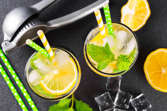 Refreshing cold icy beverage, homemade lemonade with fresh lemon slices, mint leaves, ice cubes wtih soda water, metal juicer and condensation in glasses on black textured background