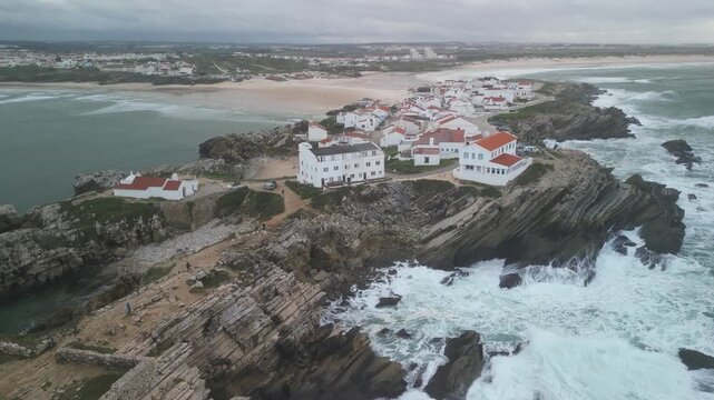 Aerial view of Baleal Island with white houses on rocky cliffs surrounded by crashing waves in Peniche, Leiria District, Portugal.