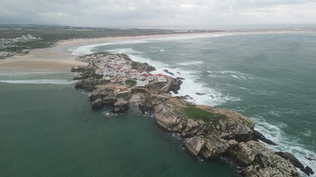 Aerial view of Baleal Island with its cluster of white houses, rocky coastline, and surrounding Atlantic Ocean under a cloudy sky in Peniche, Leiria District, Portugal.