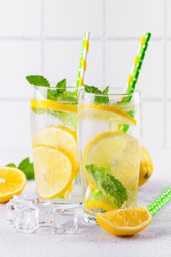 Close up of two clear glasses with refreshing homemade lemonade with fresh lemon slices, mint leaves, ice cubes and paper straws on a light kitchen counter against white tile background