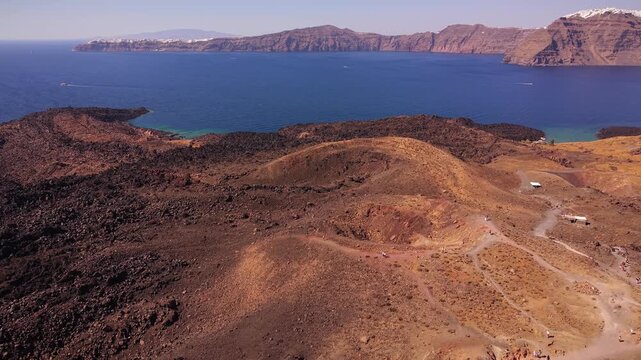 Scenic drone aerial footage hovering above the Tholos Naftilos volcanic crater in Santorini, pointing towards the iconic tourist destination of Oia, Greece.