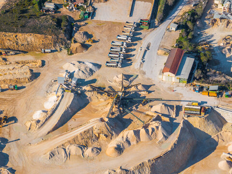 Aerial view of a stone quarry featuring piles of gravel, conveyor belts, and a line of parked dump trucks in Capestrano, Abruzzo, Italy.
