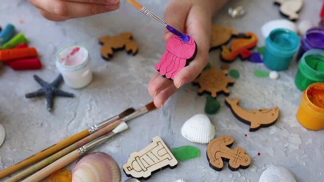 Close-up of child's hands coloring small wooden jellyfish shape with purple paint and paintbrush, surrounded by sea shells, brushes, and nautical shapes on grey background