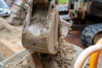 A rusted, soil-crusted mechanical digger scoop poised above a load of rubble, with the rubber tracks of the mini-excavator blurred in the background of a building zone. © Sergej