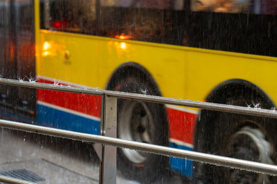 Close-up view of a metal barrier enduring heavy rainfall. The blurred wheels and colorful side panels of a large public bus provide a vibrant backdrop to the frozen water splashes.