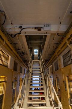Direct upward view of a long metal staircase connecting multi-story cellblock galleries with yellow walls, barred doors, and institutional architecture.