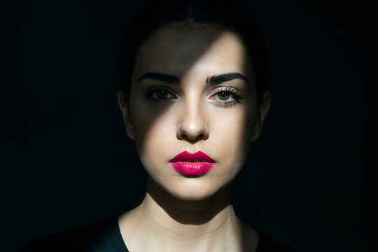Frontal view of a young woman with dark hair and vivid pink lipstick, partially obscured by a diagonal shadow against a solid black background