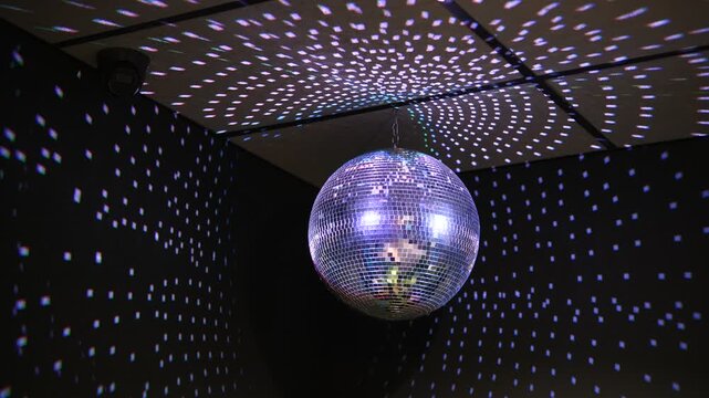 Shiny disco ball hanging from ceiling in dark room with colorful light reflections on walls. Night club party decoration with spotlight. Music dance entertainment celebration