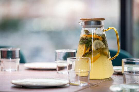 Glass pitcher of homemade lemonade with lemon, orange and mint on wooden table with empty glass and plate. Refreshing summer drink for party. Healthy food and beverage concept.