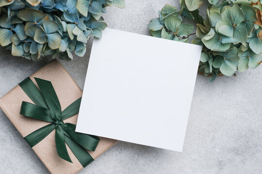 Square blank paper card placed on a gift box with a green ribbon, surrounded by dried hydrangea flowers on a textured gray surface