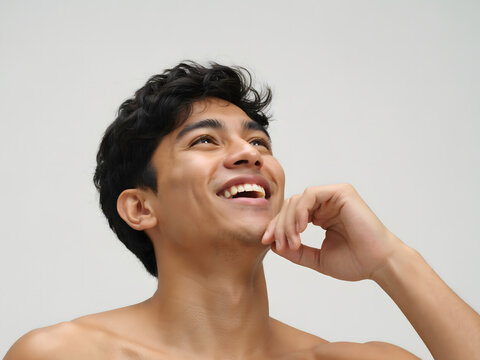 A shirtless young man with dark curly hair looking up and smiling with his hand near his chin, set against a plain light gray background