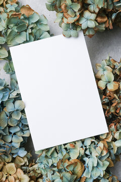 Blank wedding invitation card placed among dried blue and brown hydrangea flowers on a textured gray surface, suitable for various event announcements or personal messages