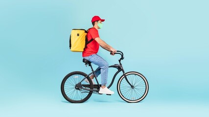 Fototapeta premium Focused Delivery Man Riding Bicycle With Yellow Delivery Bag, Wearing Face Mask, In Urban Environment. Promoting Health And Safety While Providing Delivery Services Outdoors.
