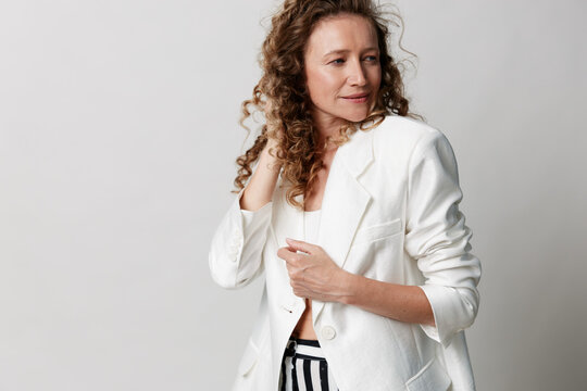 Authentic middle-aged woman with natural skin and curly hair in a white blazer posing against a light background. Studio portrait showing real beauty and confidence.