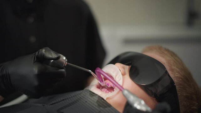 Close up of dental professional using specialized tools to examine and treat teeth in patient mouth. Dental dam protective eyewear worn by subject visible during procedure. Clinical setting