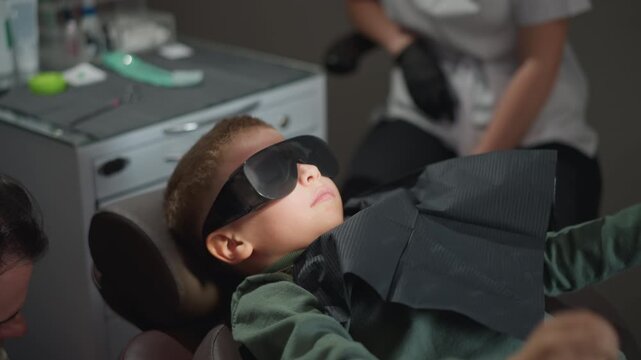 Close up of young boy lying on dental chair wearing protective eye gear and bib during orthodontic procedure. Dental assistant in background prepares tools at workstation with medical