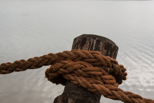 Thick rope wrapped around a weathered wooden post beside calm water. Strong textures and natural materials create a nautical scene suitable for marine, dock and craftsmanship themes.