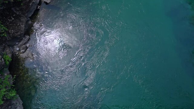 Drone view looks straight down on rippling turquoise river water swirling beside a mossy rocky bank, with sunlight glinting on the surface in a natural gorge setting.