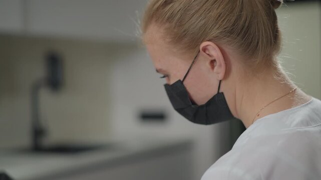 Close up of woman wearing black face mask and white shirt focused on her profile in soft lighting. background shows indoor setting with neutral tones suggesting calm concentration
