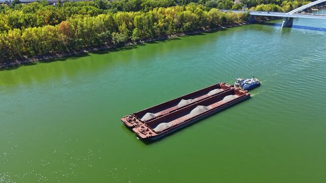 A barge moves through a river with gravel loaded on its deck. Trees line the banks of the water. The scene shows a clear day with green water.