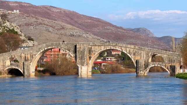 An old stone arch bridge crosses a calm river beneath rocky hills, with a small town visible along the bank in Trebinje, Bosnia and Herzegovina.