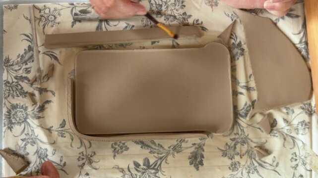 A person shapes and trims flat slabs of grey clay into a rectangular vessel form on a floral patterned cloth, an artisan pottery making process at a home studio.
