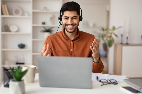 Smiling Young Caucasian Man Wearing Headset Engaging In Video Call While Working On Laptop At Home Office, Conveying Positive Emotion And Professionalism In Modern Workspace. Business Concept.