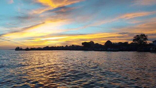 Dramatic golden sunset over Biak, Papua, Indonesia, with orange and blue clouds above a calm ocean, silhouetted coastal village, port, and ships, and warm light reflecting on rippling water.