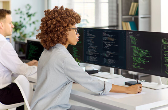 Developers programmer woman coding software in office. She writes clean code on dual monitors and computers beside a coworker. Workspace emphasizes agile teamwork. Modern tech team concept now.