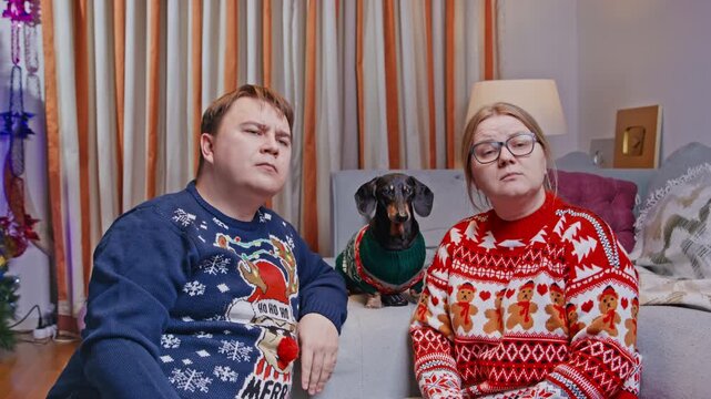 A man and woman in christmas sweaters sit on a sofa with a black and tan dachshund dog in a matching sweater between them, all looking tired and unimpressed at home.