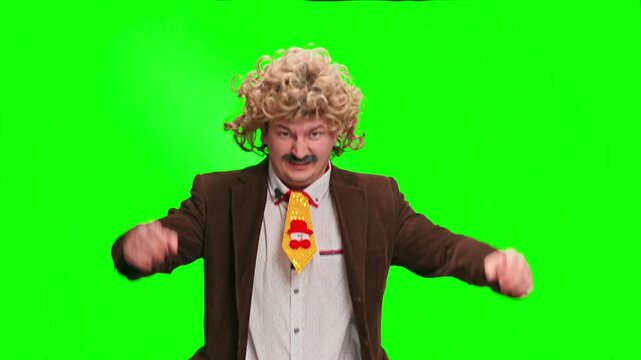 A man wearing a curly blonde wig, fake mustache and brown jacket dances and points at the camera against a green screen background, a playful comedic presenter setup with copy space.