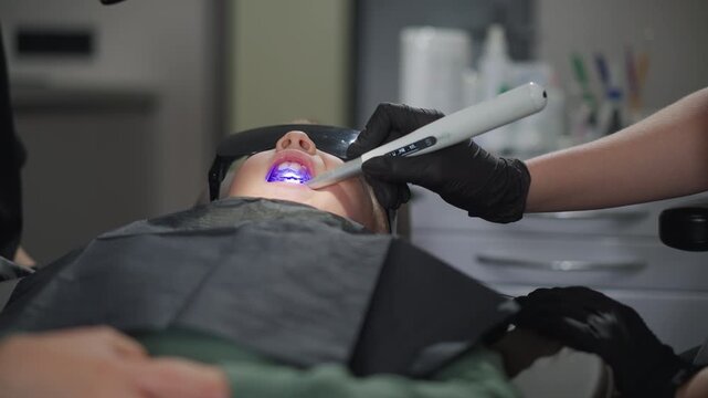 Close up of young woman lying on examination chair while undergoing teeth whitening treatment. dental professional wearing black gloves uses handheld device to apply and activate bleaching