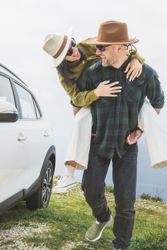 Close up happy couple enjoys holiday time together piggyback check mirror by white SUV during Mediterranean road trip. Lifestyle shot capturing love, freedom adventure on scenic coastal cliff side