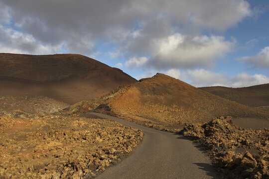 Asphalted road in national park, red-brown lava landscape, Timanfaya National Park, fire mountains, volcanic landscape, blue sky, grey-white clouds, Lanzarote, Canary Islands, Spain