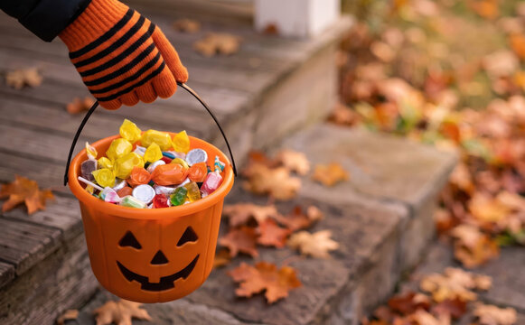 Child holding a pumpkin bucket full of candy for trick or treat on Halloween with autumn leaves