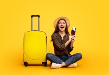 Smiling Young Woman With Light Skin And Brown Hair Holding Passport And Ticket While Sitting On Floor Beside Yellow Suitcase. Happy Emotion Reflects Travel Enthusiasm In Studio Setting. © Prostock-studio