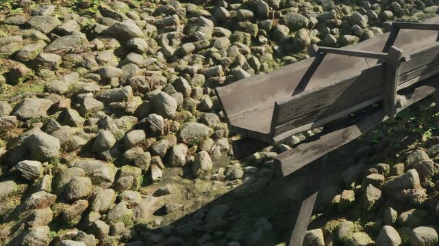 A wooden structure stands near a rocky streambed covered with various sized stones. Sunlight shines on the scene, revealing patches of green moss and small plants.