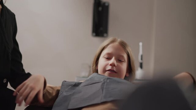 Close up of young girl lying on examination chair in dental office while her teeth are being worked on. girl adjusts patient bib and positions their hand near with soft lighting illuminating