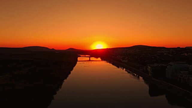 The sun sets over a river with mountains on one side and buildings on the other. The sky shows orange and yellow colors while the water reflects the scene below.