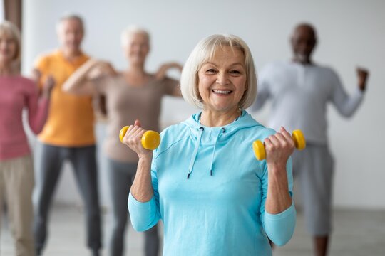 Smiling Senior Woman Holding Dumbbells While Exercising With Group Of Senior Friends, Engaging In Fitness Activity Together In Bright Gym Setting. Promoting Health And Wellness.
