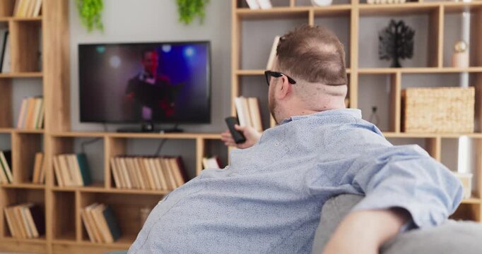 Overweight man relaxing at home, watching tv. He sits on a sofa, an adult, using a remote to change channels and stream shows in a living room at home. Strong concept of home media relaxation.