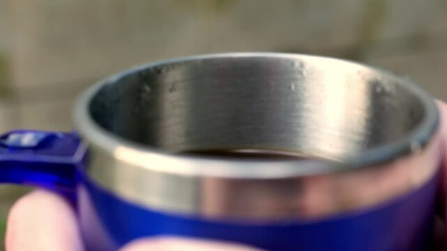 POV of a man holding a steaming cup of black coffee in the sunlight. Close-up: man holding an espresso mug as he drinks coffee. Drinking coffee time. An insulated tumbler. A mug of coffee in the sun. 
