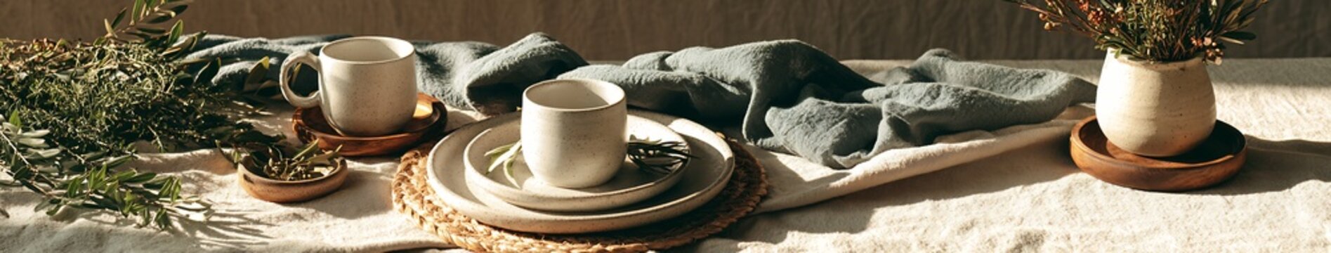 Winter Holiday Rustic neutral table setting with speckled ceramic plates and mugs, greenery and woven placemat in warm natural light